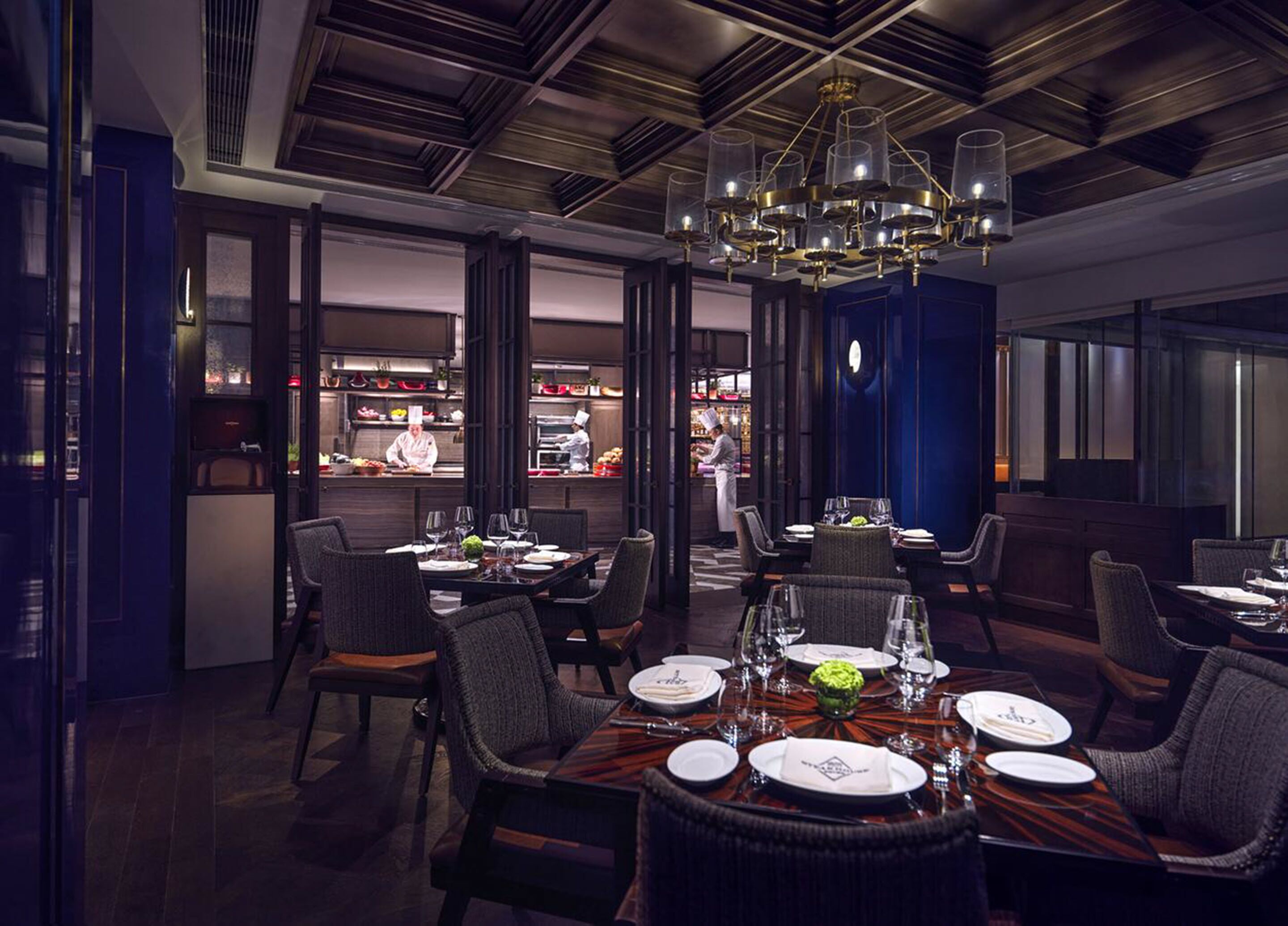 A restaurant with a dark wood ceiling, a chandelier, and a view of the kitchen.
