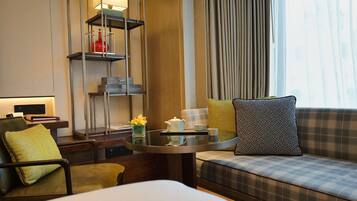 A hotel room with a bed, a sofa, a small table with a teapot and cups, a bookshelf, and a window with curtains.