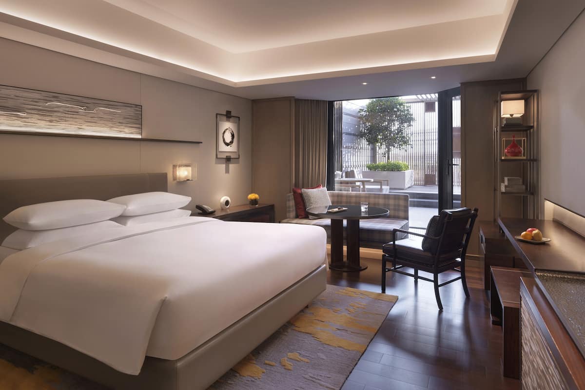 A modern hotel room with a large bed, a desk with a chair, a small table, and a view of the outdoors.