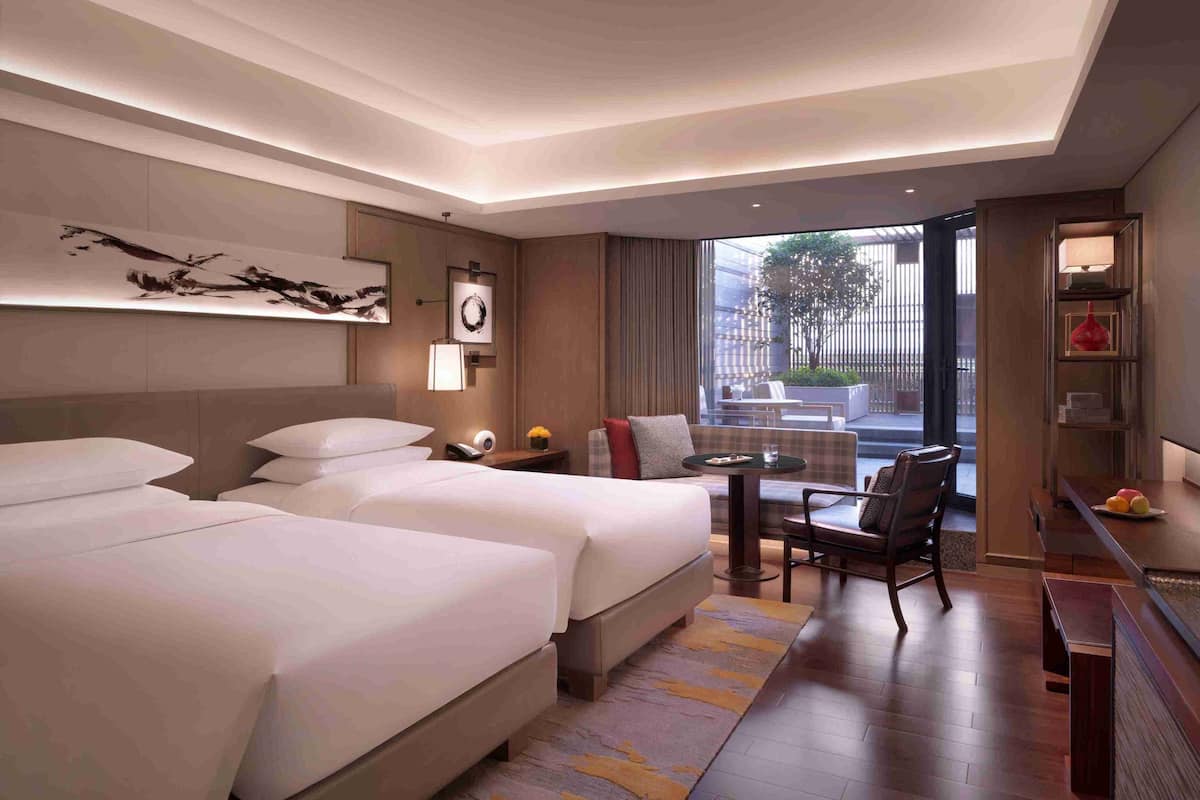 A hotel room with two beds, a desk, a chair, a window with a view, and a wall-mounted artwork.
