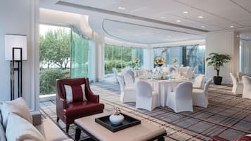 A modern hotel lobby with a seating area, a dining setup, and large windows offering a view of greenery.