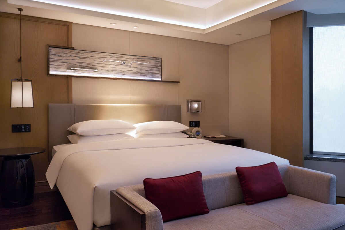A modern hotel room with a large bed, a bedside table, a nightstand, a wall-mounted artwork, and a window providing natural light.