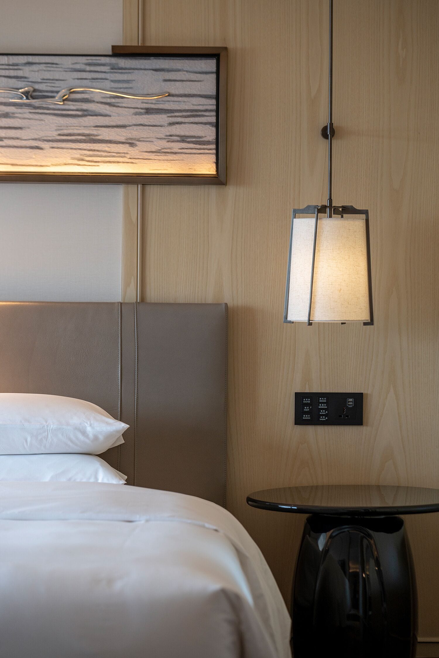 A hotel room with a bed, a bedside table, a lamp, a wall-mounted control panel, and a painting.
