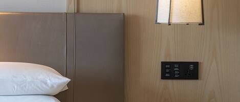 A hotel room with a bed, a bedside table, a lamp, a wall-mounted control panel, and a painting.