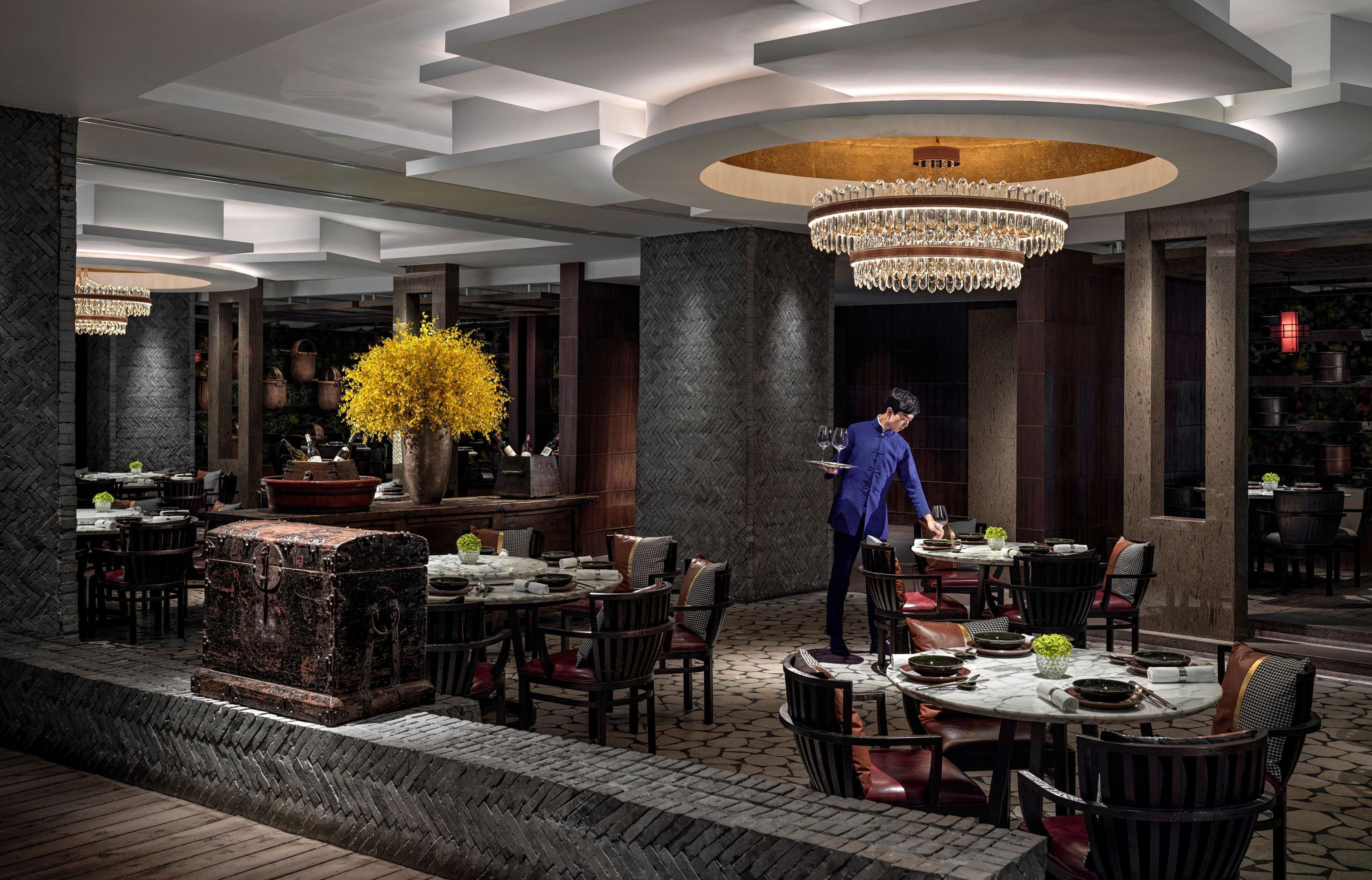 A restaurant with a central chandelier, wooden tables, and chairs.
