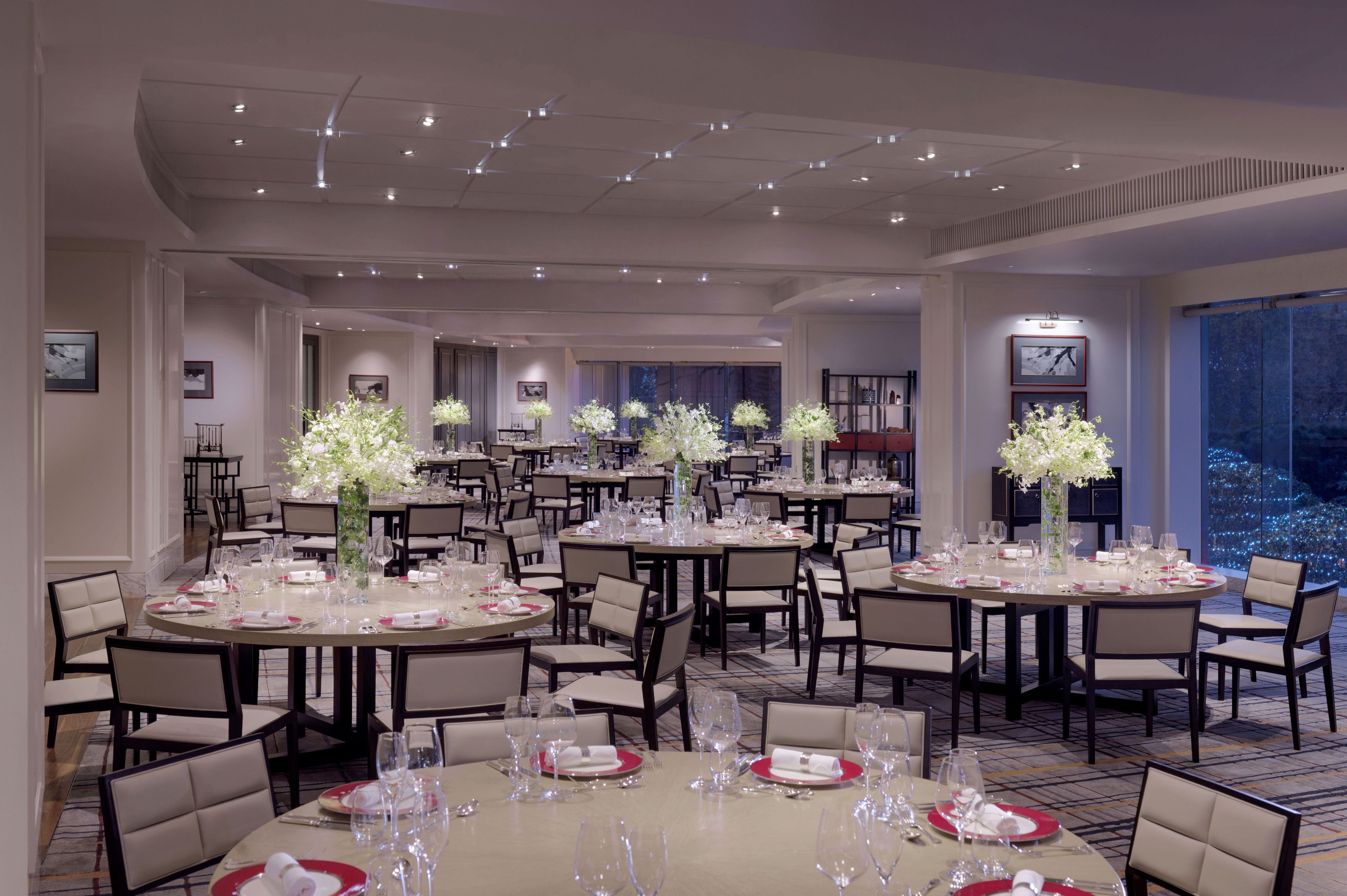 Un vaste espace dédié à la salle à manger, avec plusieurs tables rondes dressées pour le repas, chacune agrémentée d’un centre de table floral blanc et d’une décoration de table soignée.