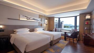 A hotel room with two beds, a desk, a chair, and a large window offering a view of buildings and mountains.