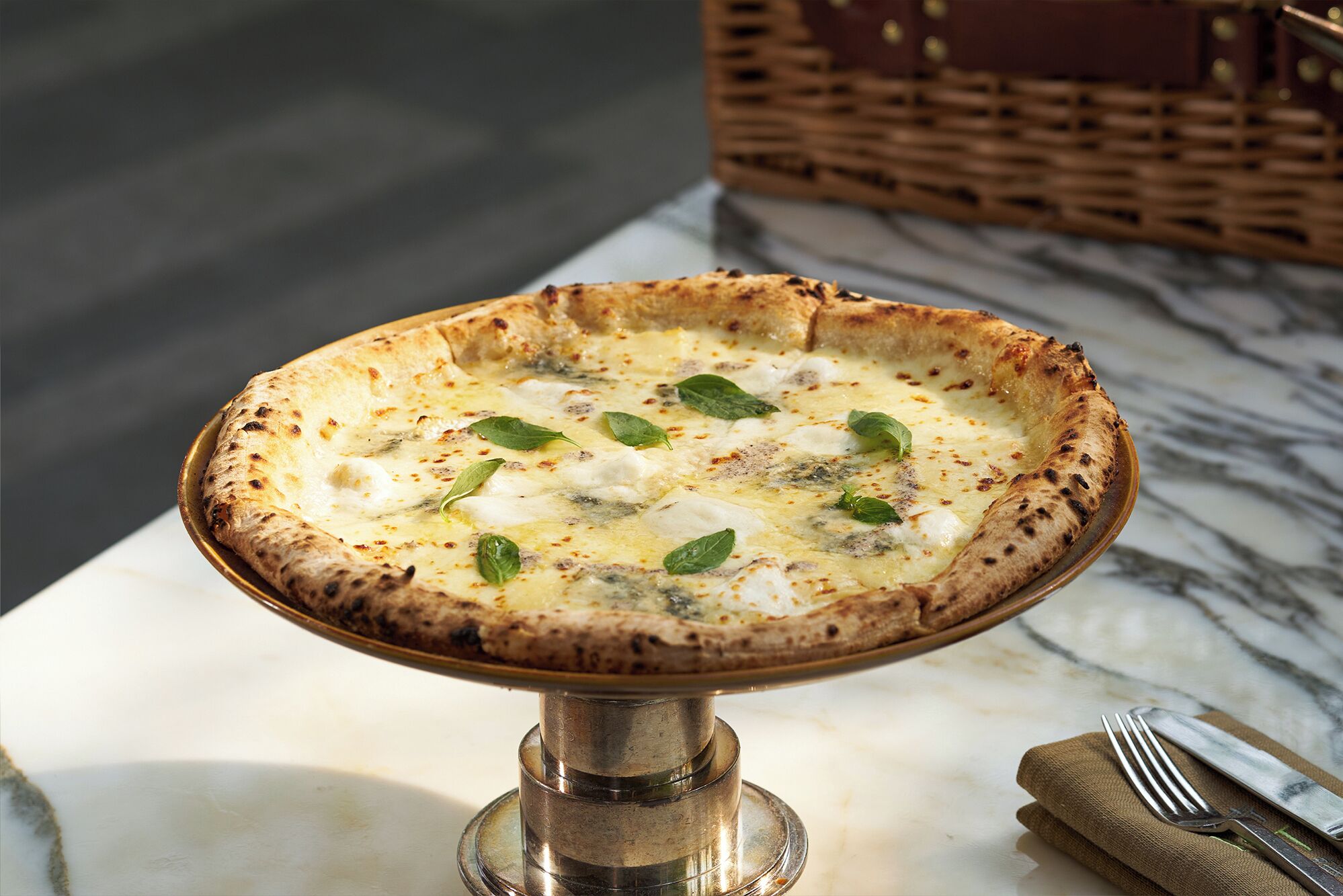 Una pizza sobre un soporte metálico, con una mesa de mármol y una cesta de mimbre al fondo.