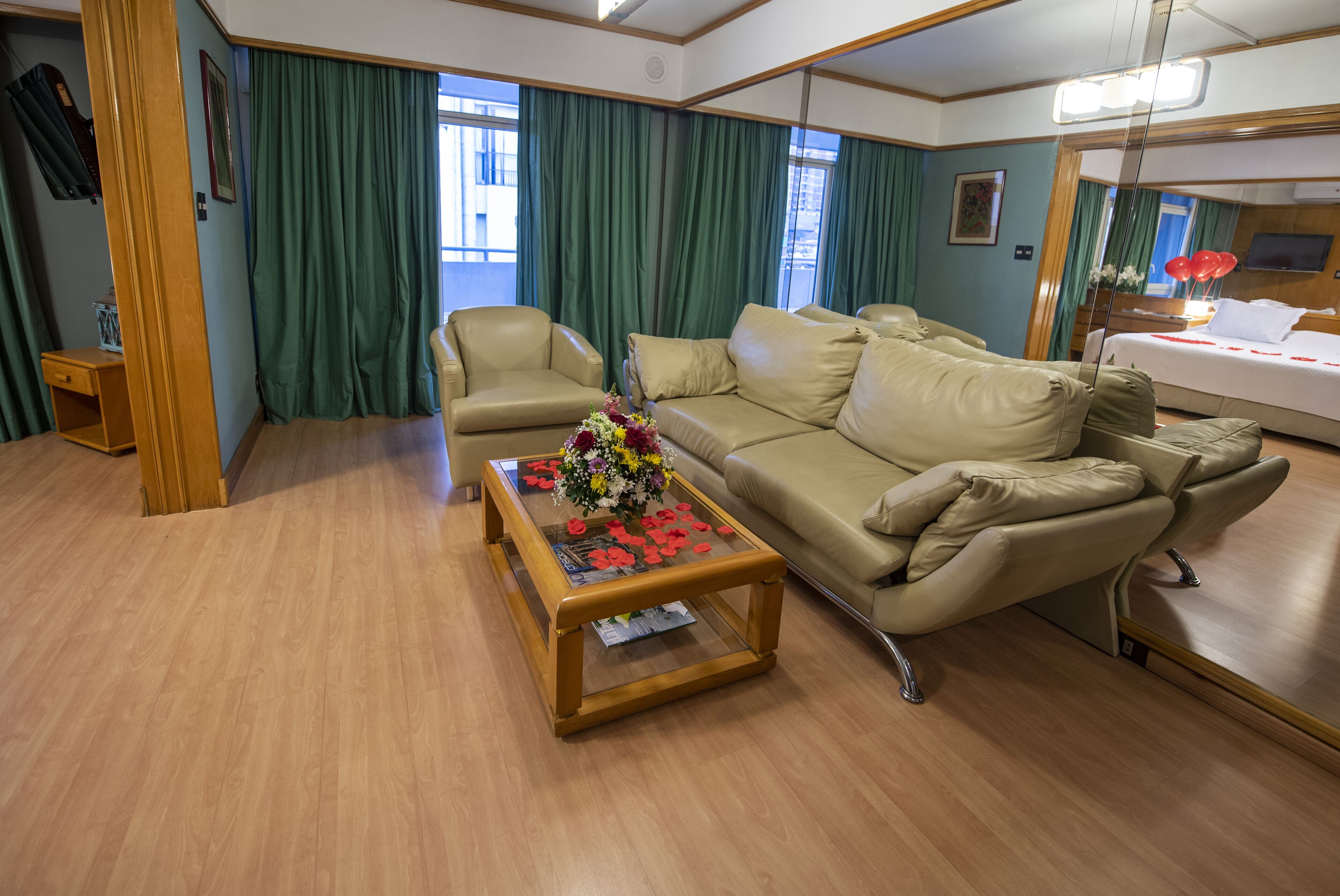 Une chambre d’hôtel avec un canapé beige, une table basse en verre agrémentée d’un arrangement floral et des rideaux verts.