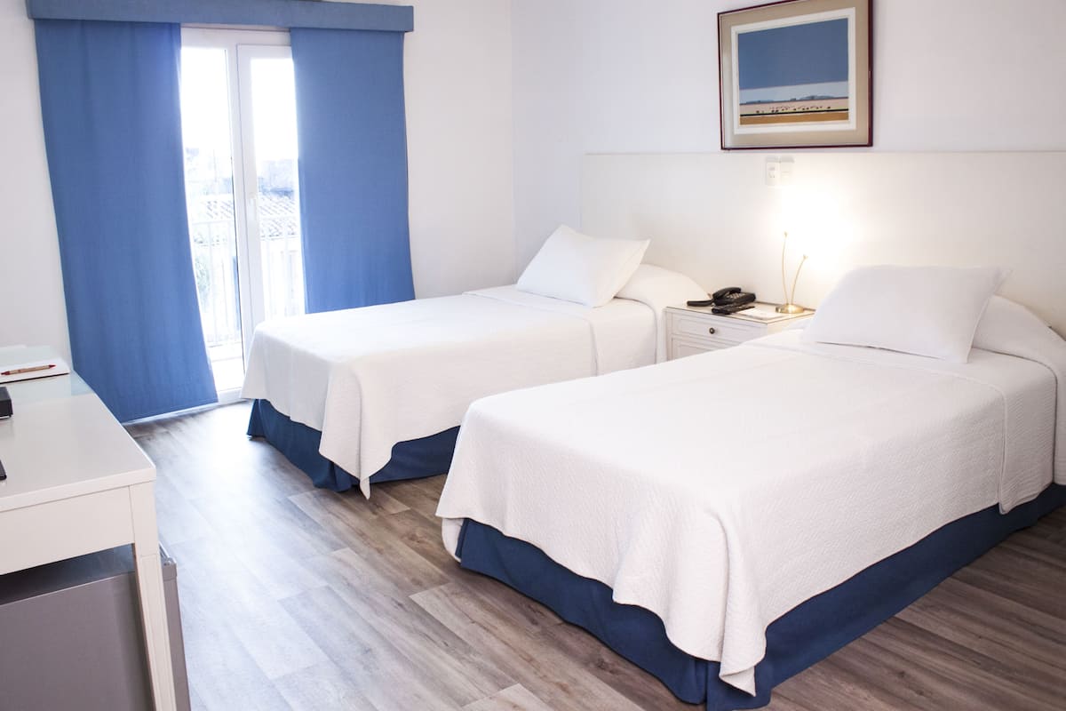 A hotel room with two single beds, a desk, and a window with blue curtains.
