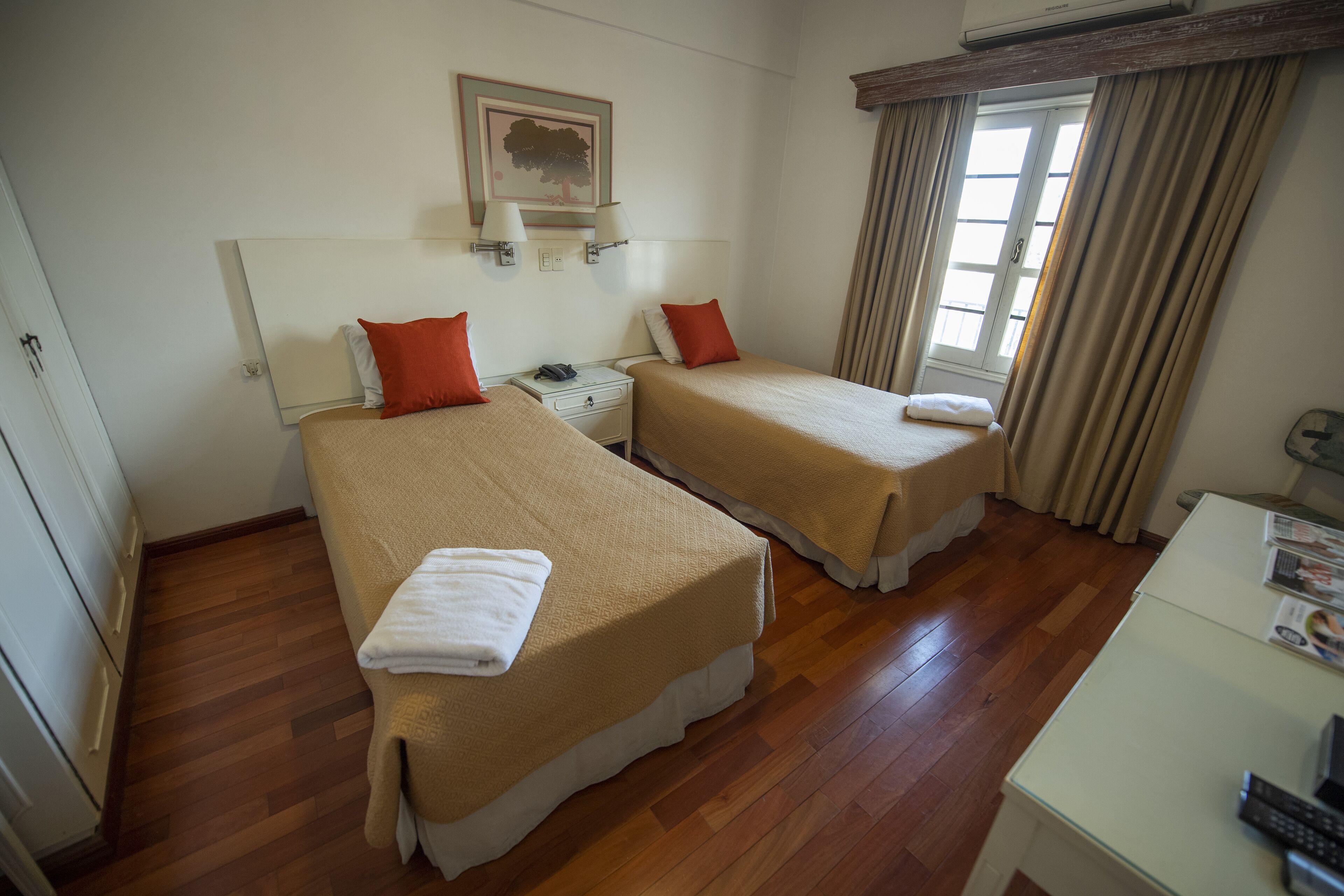 A hotel room with two beds, wooden flooring, a white desk, and a window with curtains.