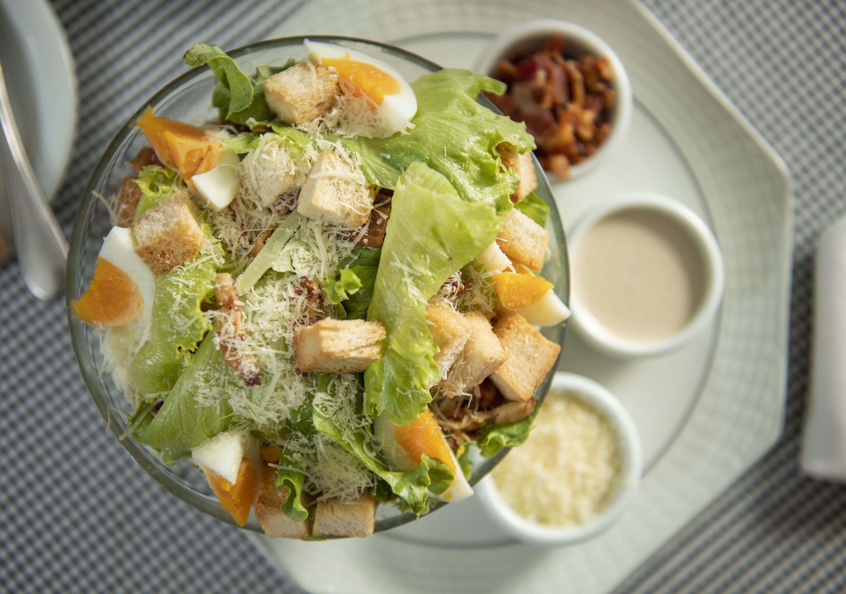 Vista chiara di una ciotola di insalata con crostini, formaggio grattugiato e un uovo sodo, accompagnata da due piccole ciotoline di condimento.
