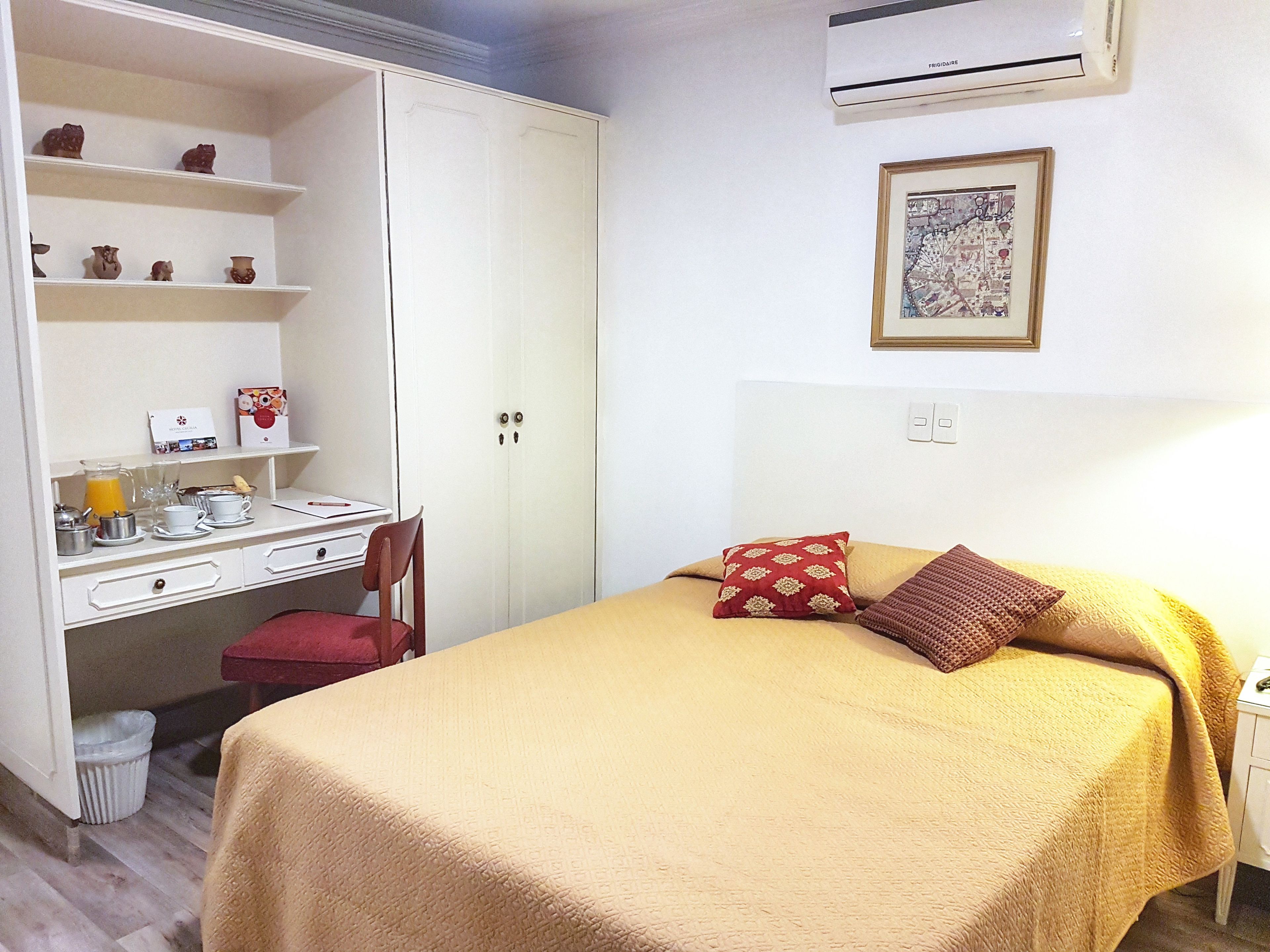 A hotel room with a bed, a desk with a chair, a wardrobe, and a small shelf with decorative items.