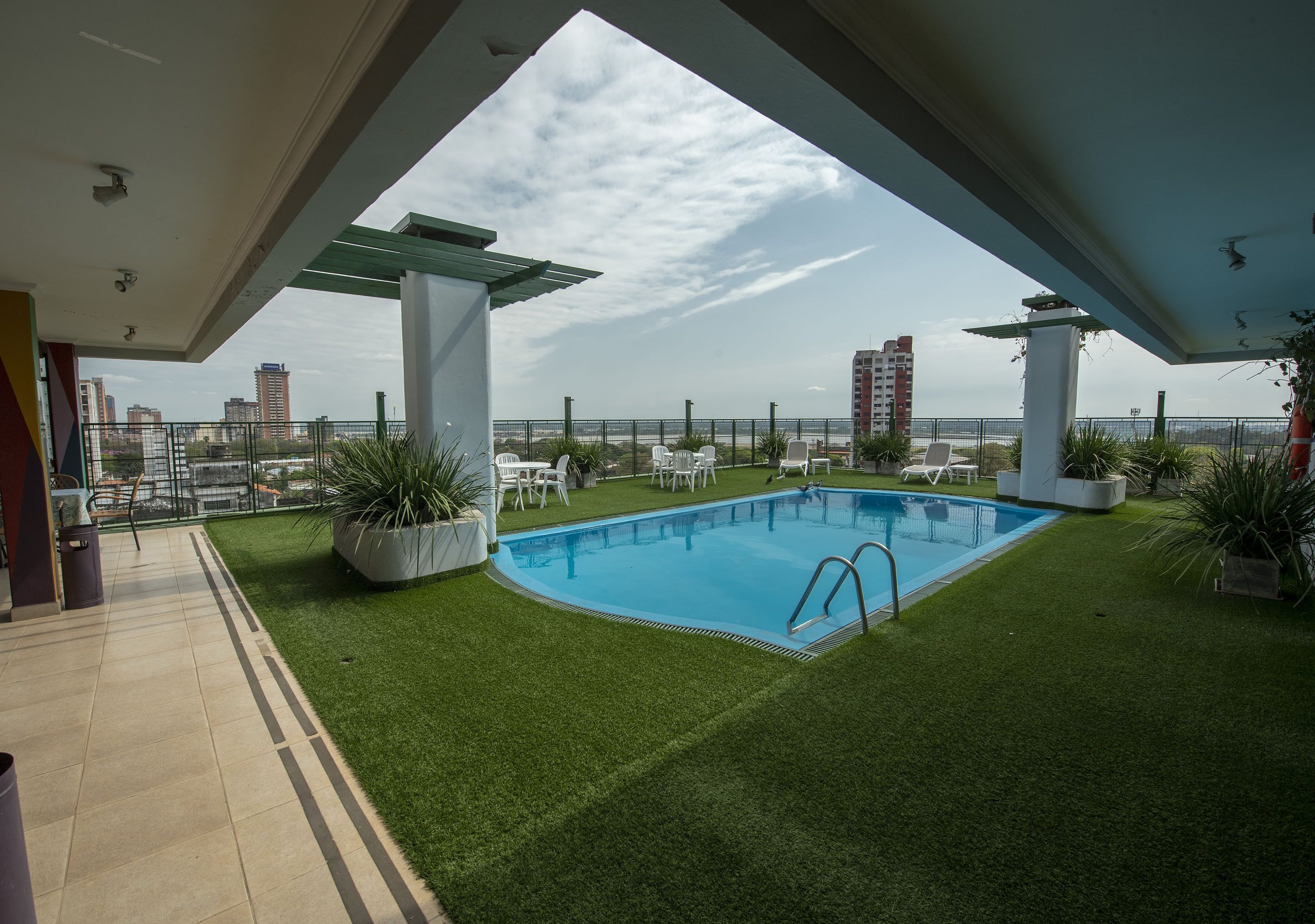 Una piscina en la azotea con césped artificial, sillas reclinables blancas y vistas al horizonte de la ciudad.