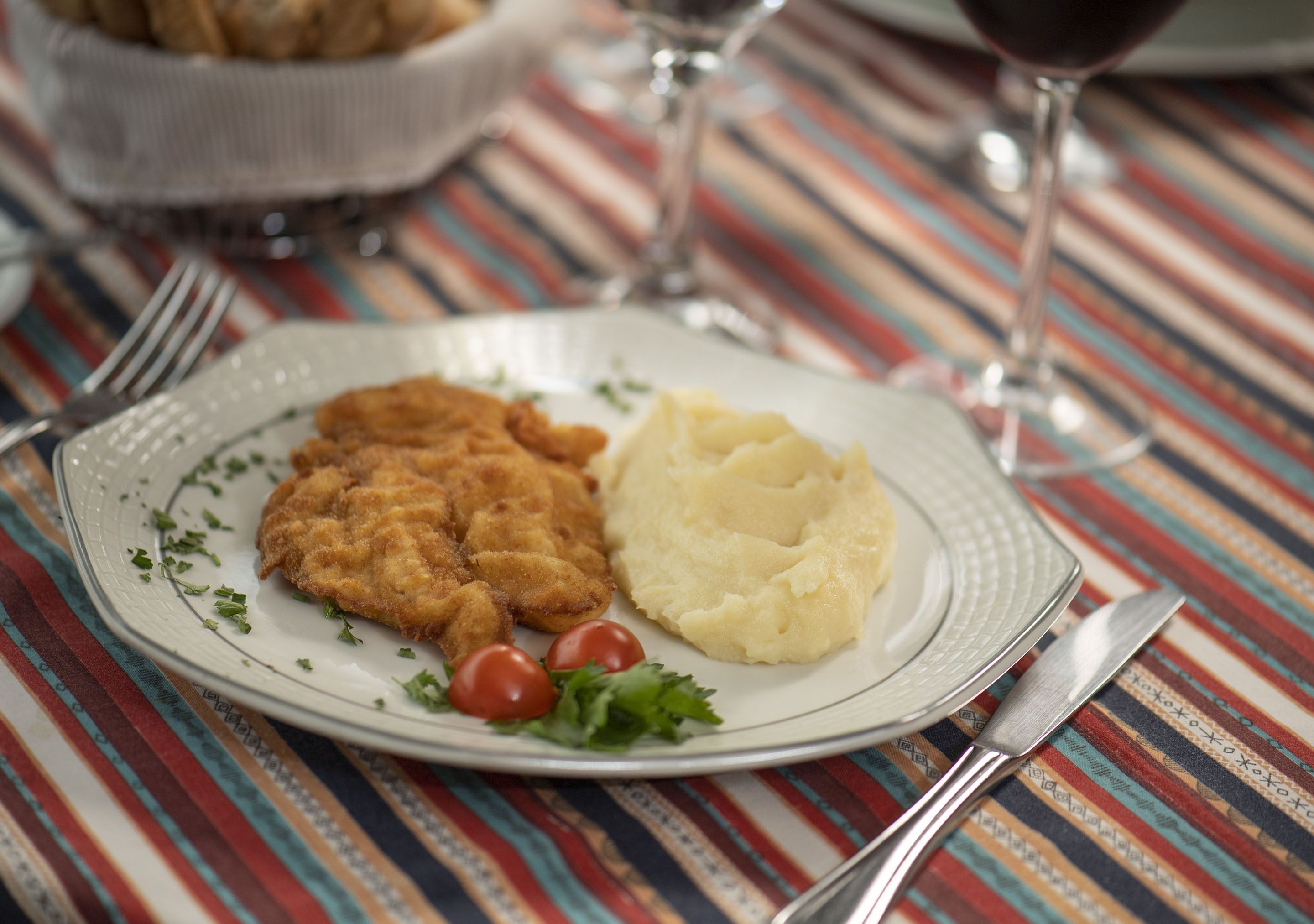 A plate with breaded meat, mashed potatoes, and cherry tomatoes on a striped tablecloth.