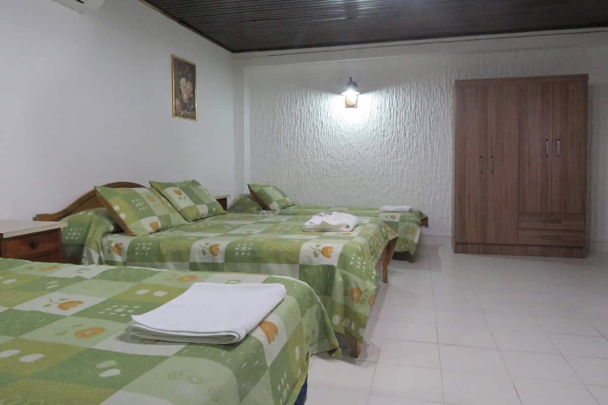 A hotel room with three beds, each with green floral bedding and white towels. There is a wooden wardrobe and a wall-mounted light fixture.