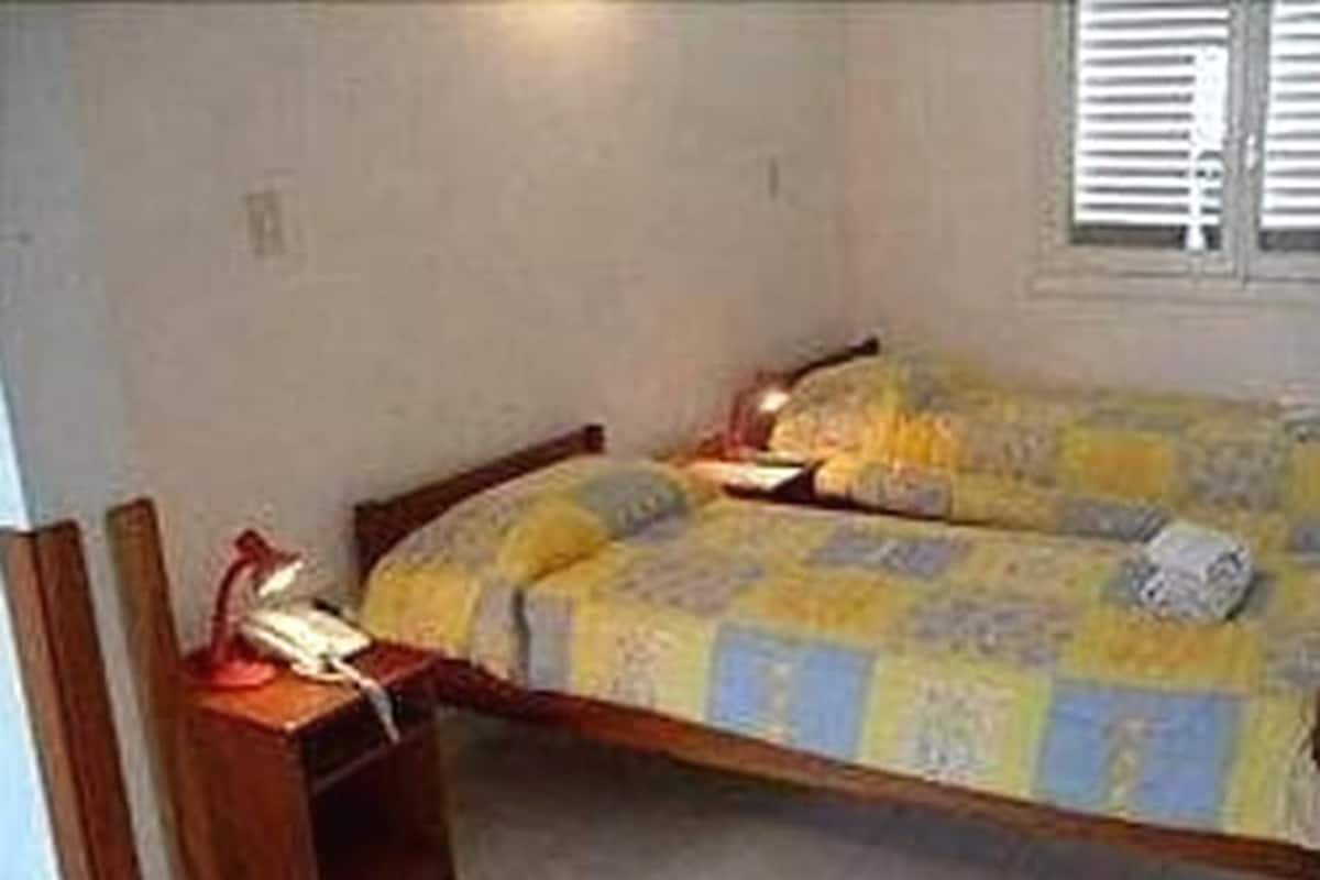 A small, single-bed room with a wooden bed frame, a bedside table with a lamp, and a window with blinds.
