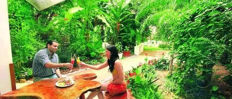 A man and a woman sitting at a wooden table in a lush garden with dense foliage and a variety of plants.