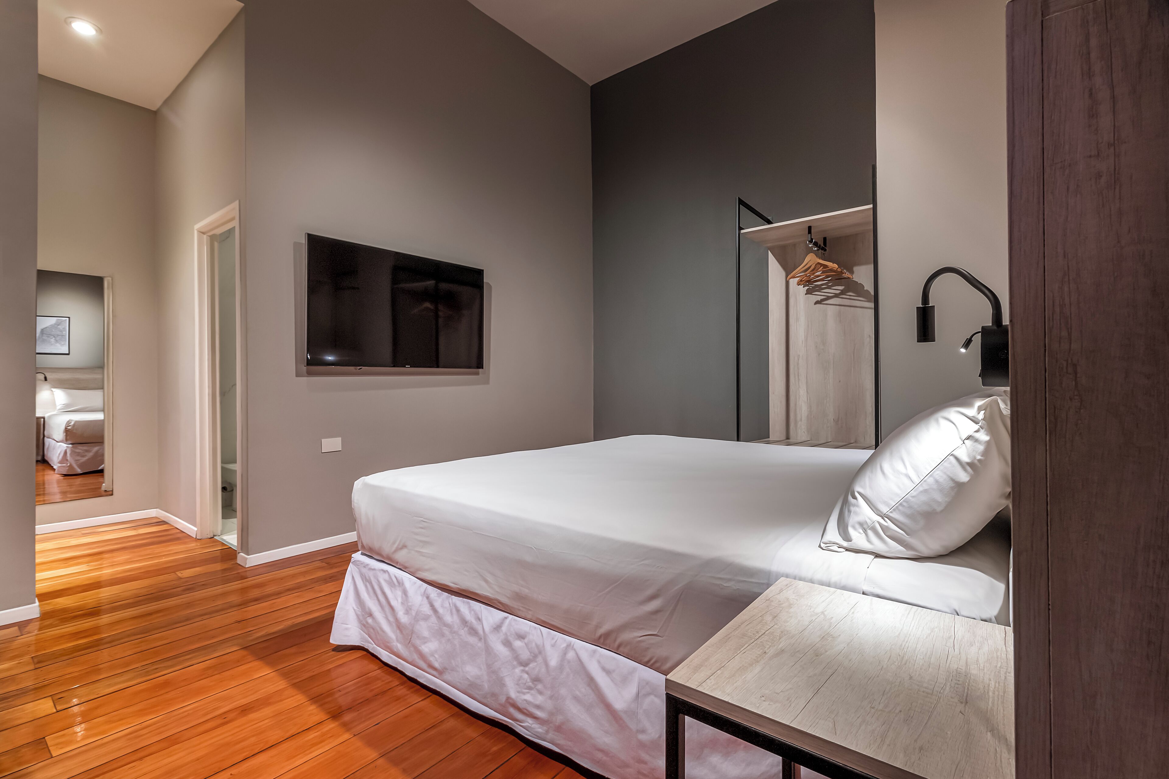 A bedroom with a bed, a bedside table, a mirror, a hanging wardrobe, and a door leading to another room.