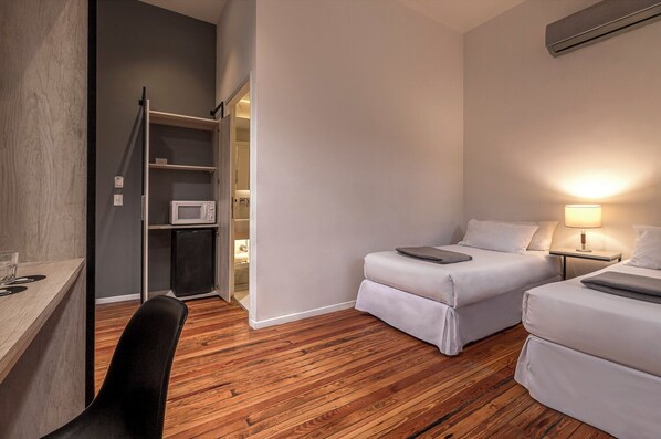 Une chambre d’hôtel compacte avec un plancher en bois, deux lits simples, un bureau, une chaise, un four à micro-ondes et un petit réfrigérateur.