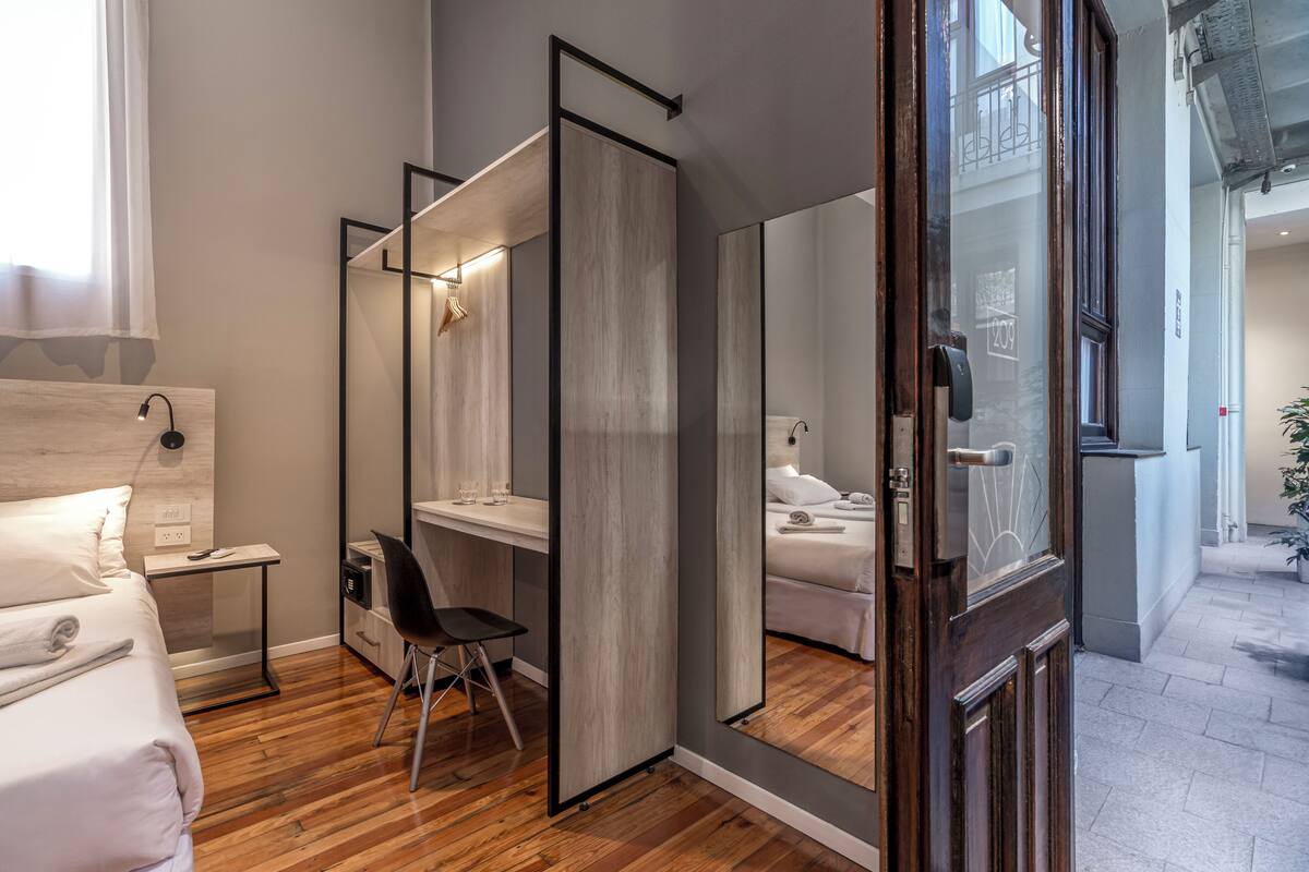 Habitación de hotel moderna con piso de madera, cama con sábanas blancas, escritorio con silla, un espejo grande y baño visible a través de un tabique de vidrio.