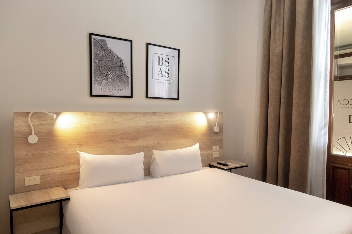 A hotel room with a wooden headboard, two white pillows, a nightstand, a framed map, and a framed sign.