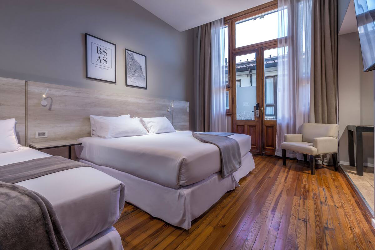 Habitación de hotel con dos camas, piso de madera, una silla y un ventanal con cortinas.