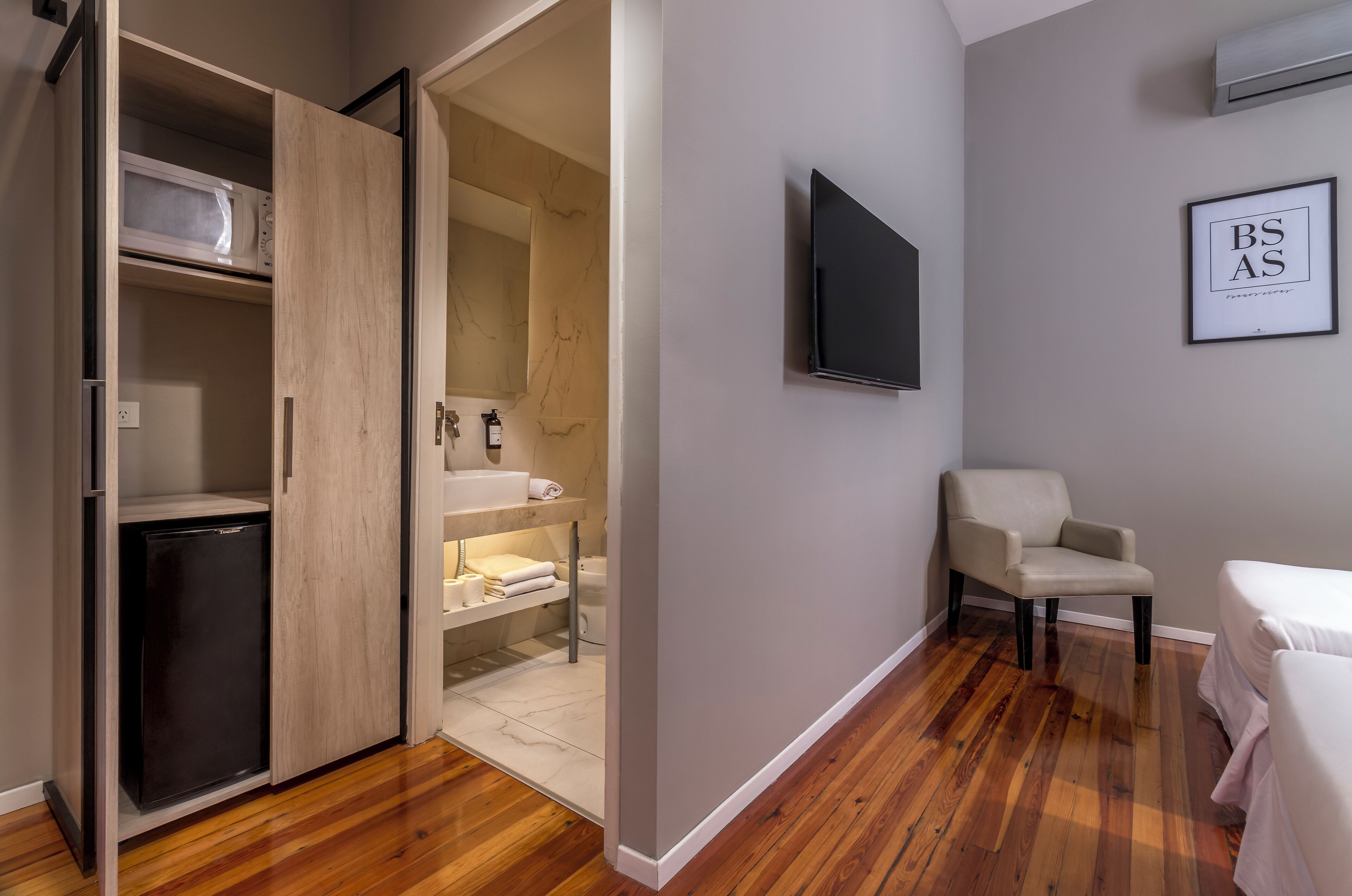 A compact hotel room with a wooden floor, a built-in wardrobe, a small refrigerator, a sink, a chair, and a bed.