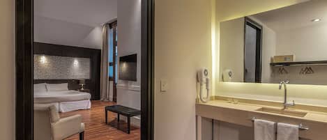 Una habitación de hotel moderna con piso de madera, una cama, un sofá, una mesita, un baño con lavamanos, un espejo y toallas.