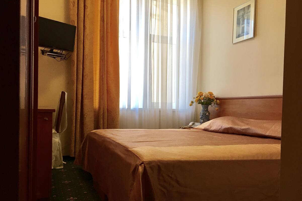 A hotel room with a bed, a nightstand with a vase of flowers, a television mounted on the wall, and a door leading to another room.
