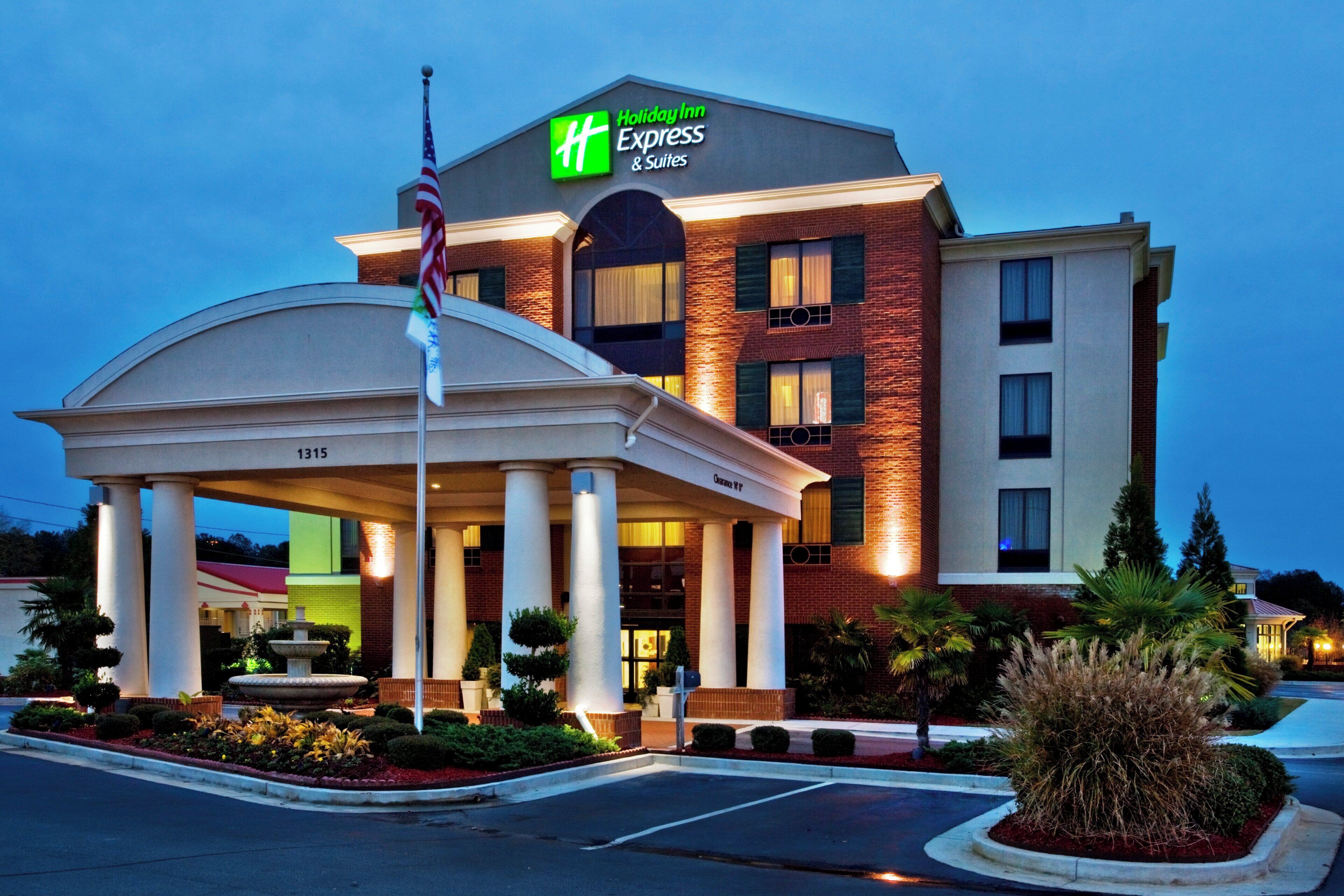 A Holiday Inn Express & Suites building with a prominent American flag, a fountain, and a well-lit entrance.