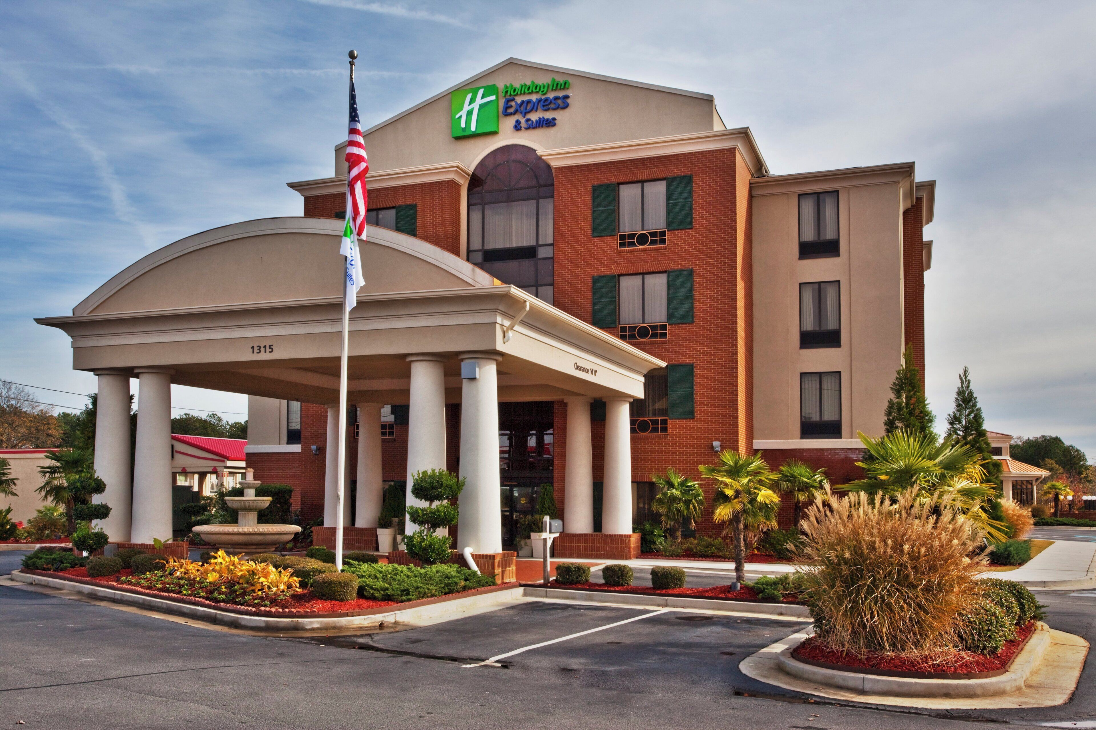 Edificio de Holiday Inn Express & Suites con entrada cubierta, asta con banderas y área ajardinada con plantas y una fuente.