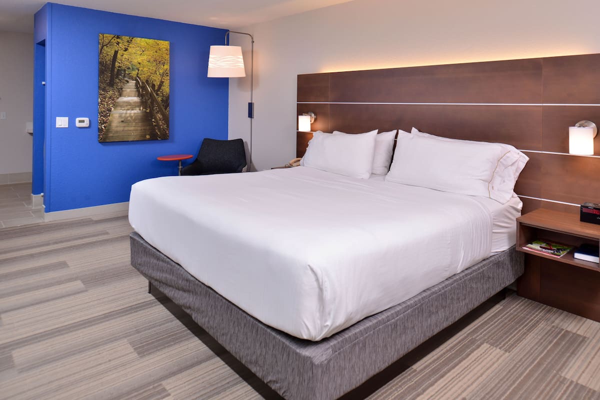 A hotel room with a large bed, a blue accent wall, a painting, and a nightstand with a lamp.