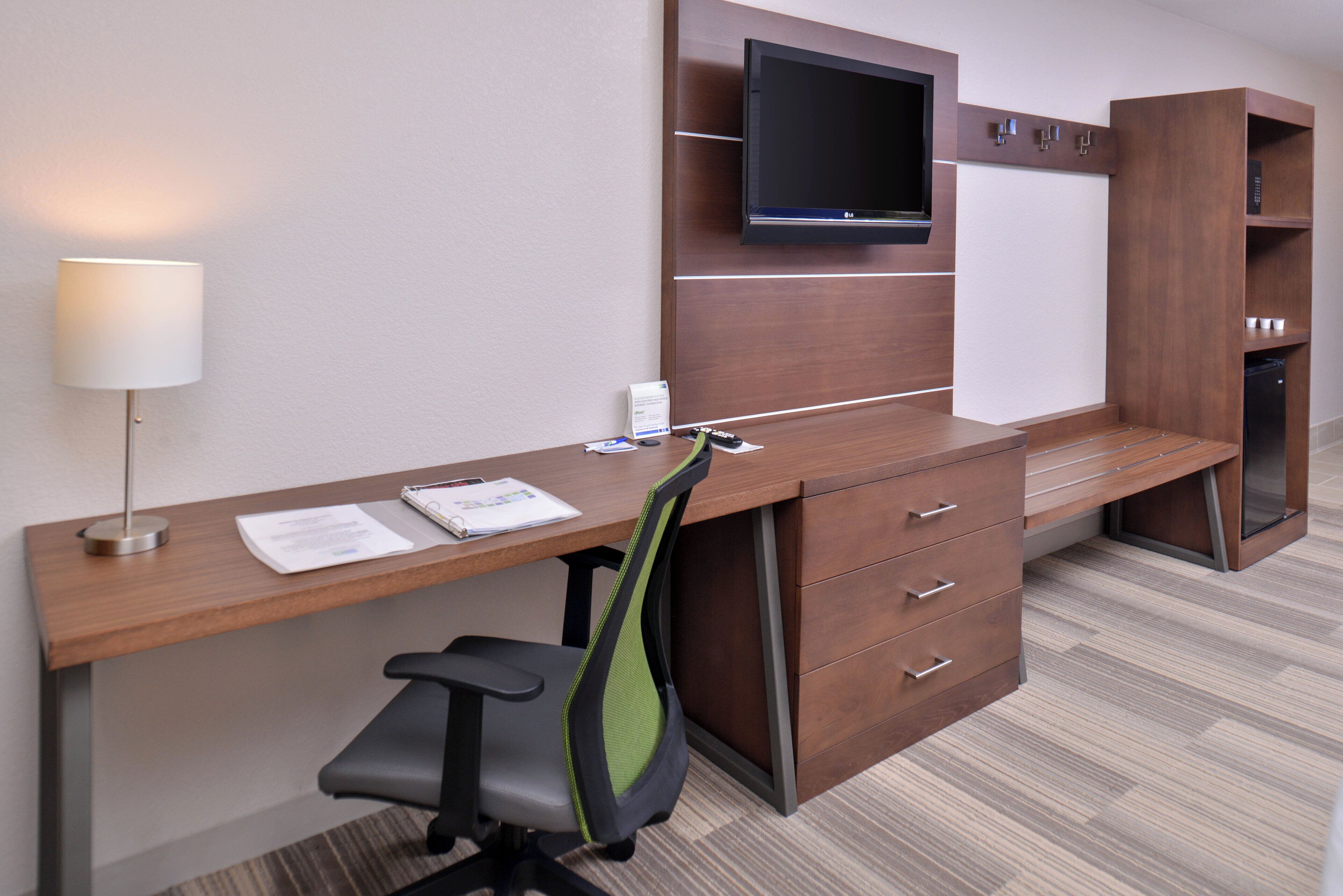 A hotel room with a wooden desk, a chair, a flat-screen TV mounted on the wall, a lamp, and a minibar.