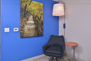 Una habitación con una pared azul, un cuadro con un sendero de madera, una silla moderna, una pequeña mesa redonda y una lámpara de pie.