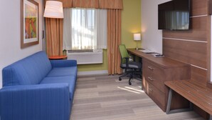 A hotel room with a blue sofa, a desk with drawers, a television mounted on the wall, a chair, and a window with curtains. - Holiday Inn Express and Suites Stevens Point by IHG (Stevens Point)