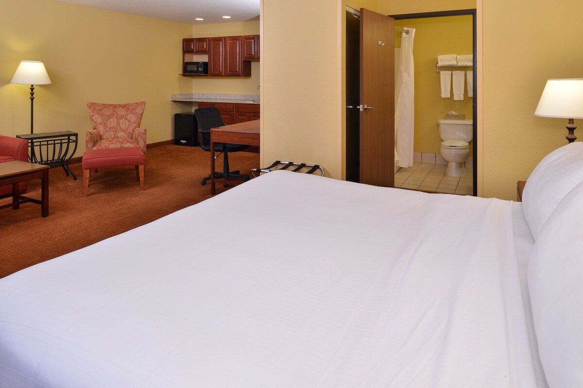 A hotel room with a large bed, a desk, a chair, and a kitchenette.