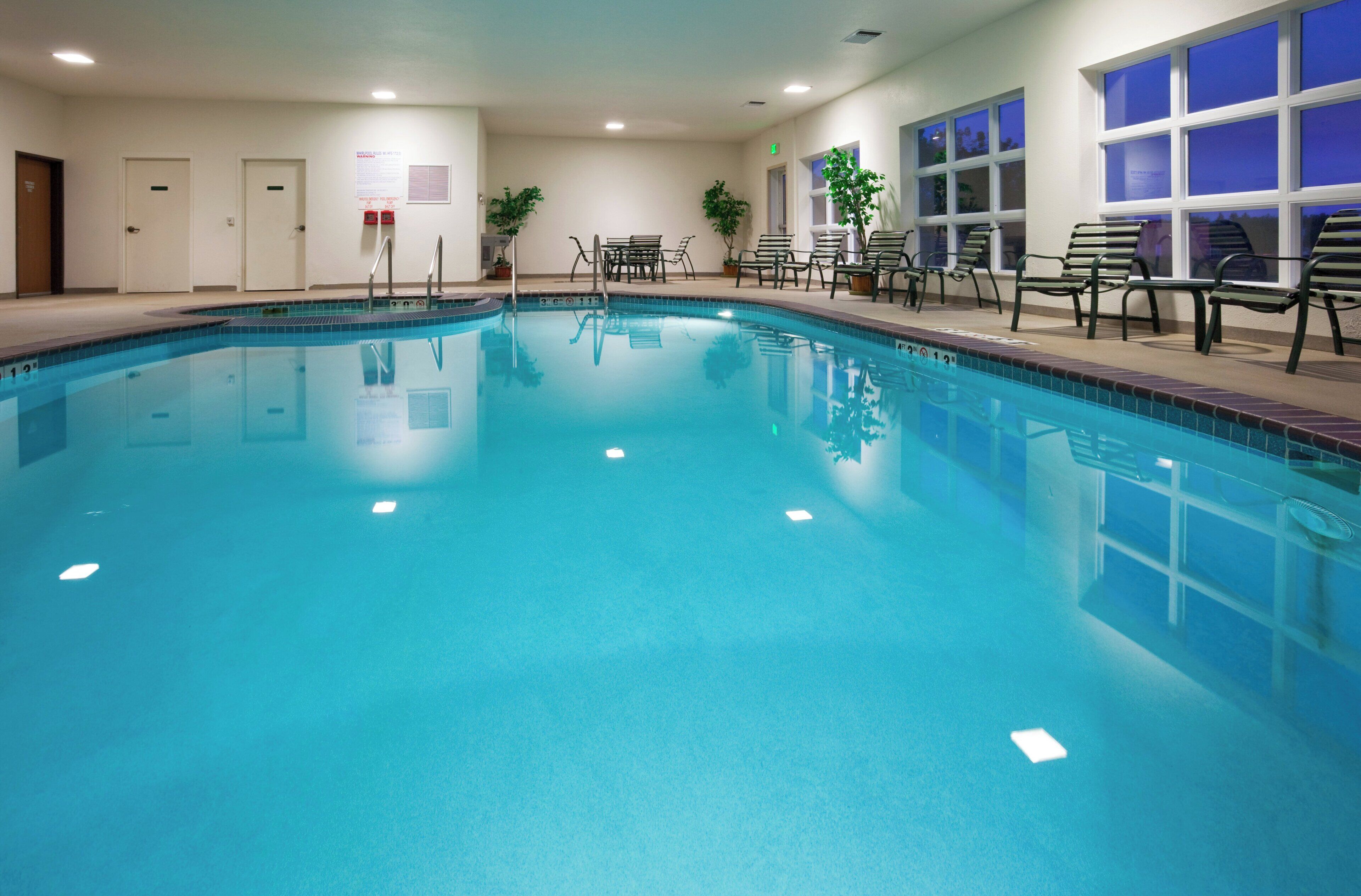 Una piscina interior con agua cristalina, rodeada de sillones de descanso y un banco, con puertas y una ventana al fondo.