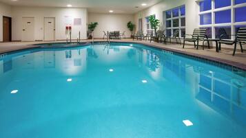 Una piscina interior con agua cristalina, rodeada de sillones de descanso y un banco, con puertas y una ventana al fondo.