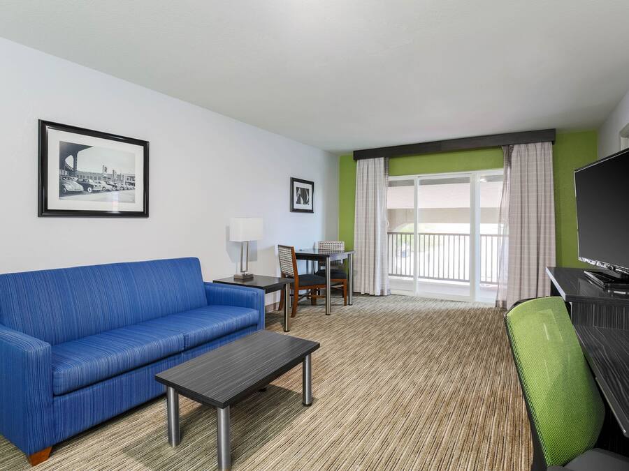 A hotel room with a blue sofa, a small table, a green chair, a TV, and a balcony with curtains.