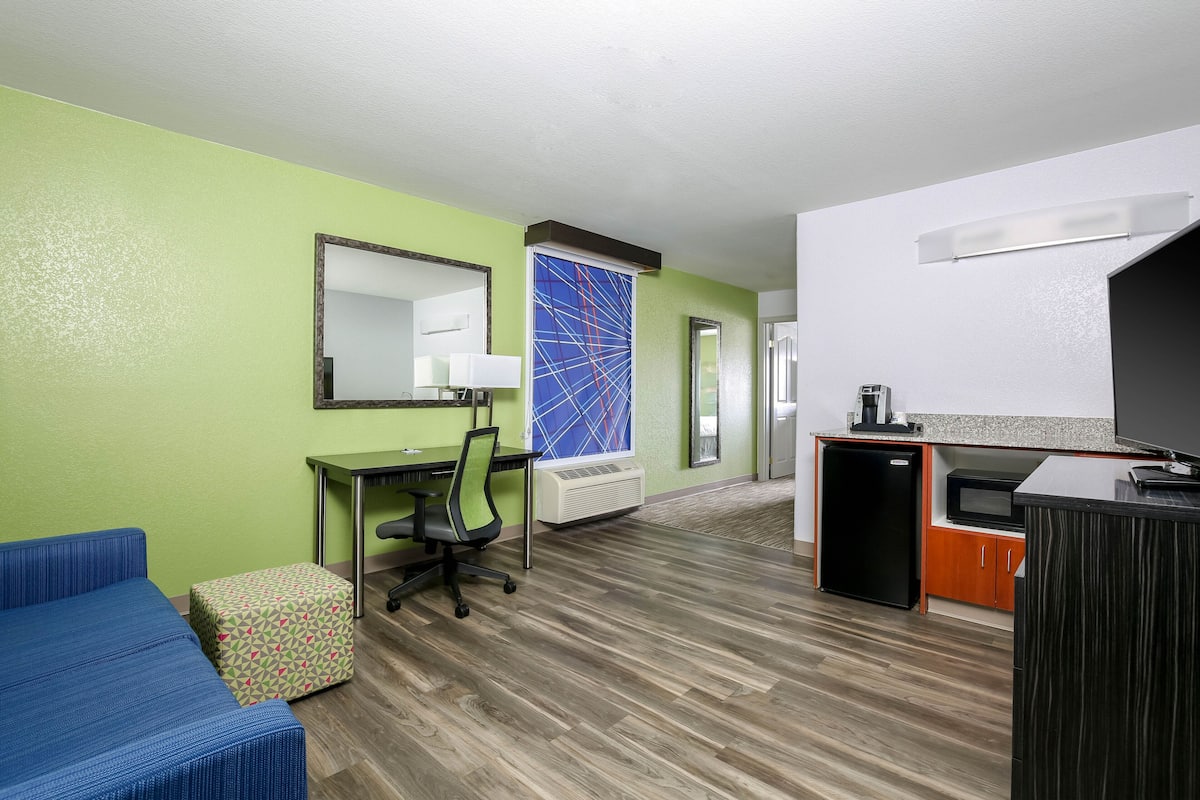 Habitación de hotel con pared verde, sofá azul, escritorio con silla, kitchenette pequeño y televisor.