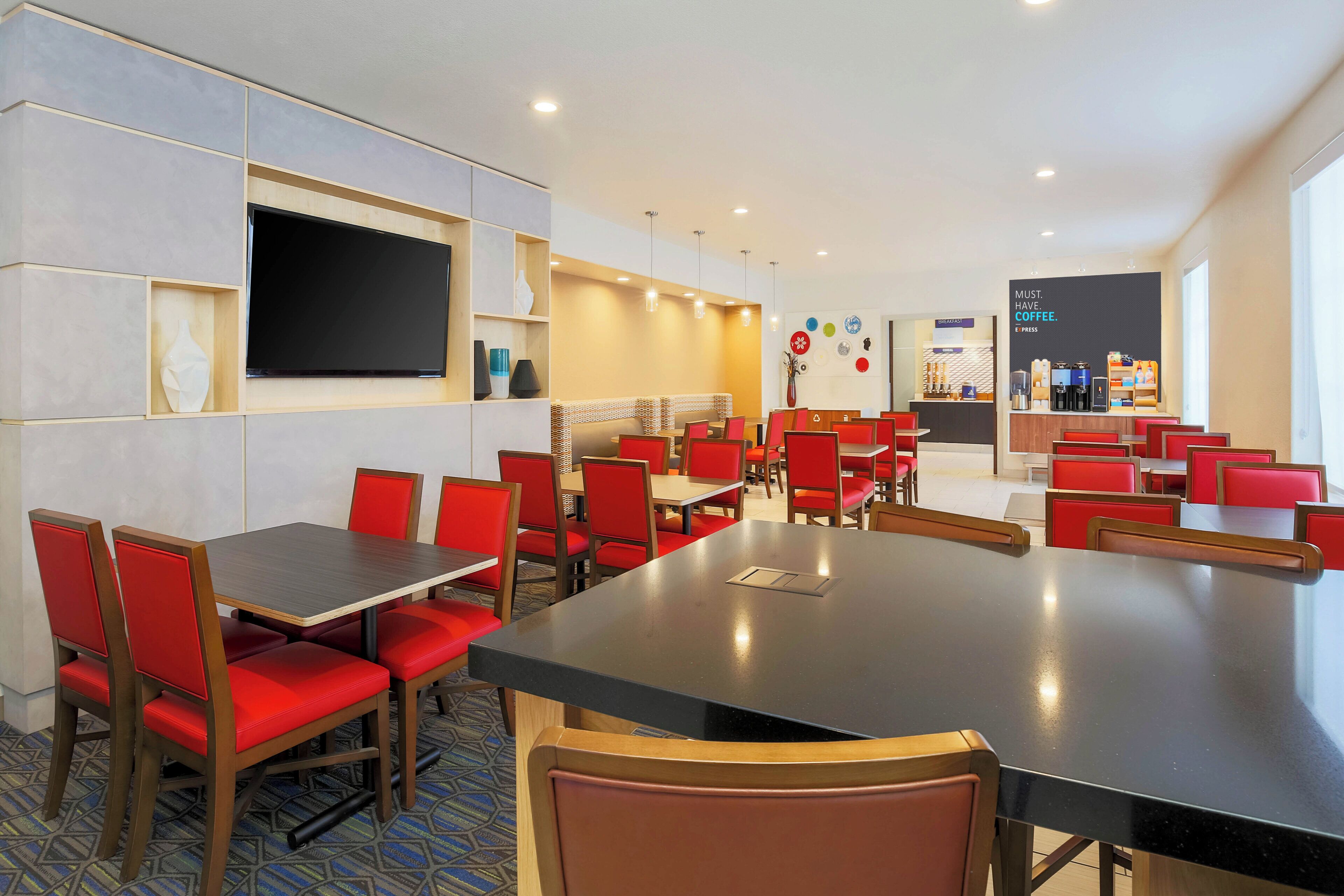 A hotel dining area with multiple tables and red chairs, a flat-screen TV mounted on the wall, and a coffee bar in the background.