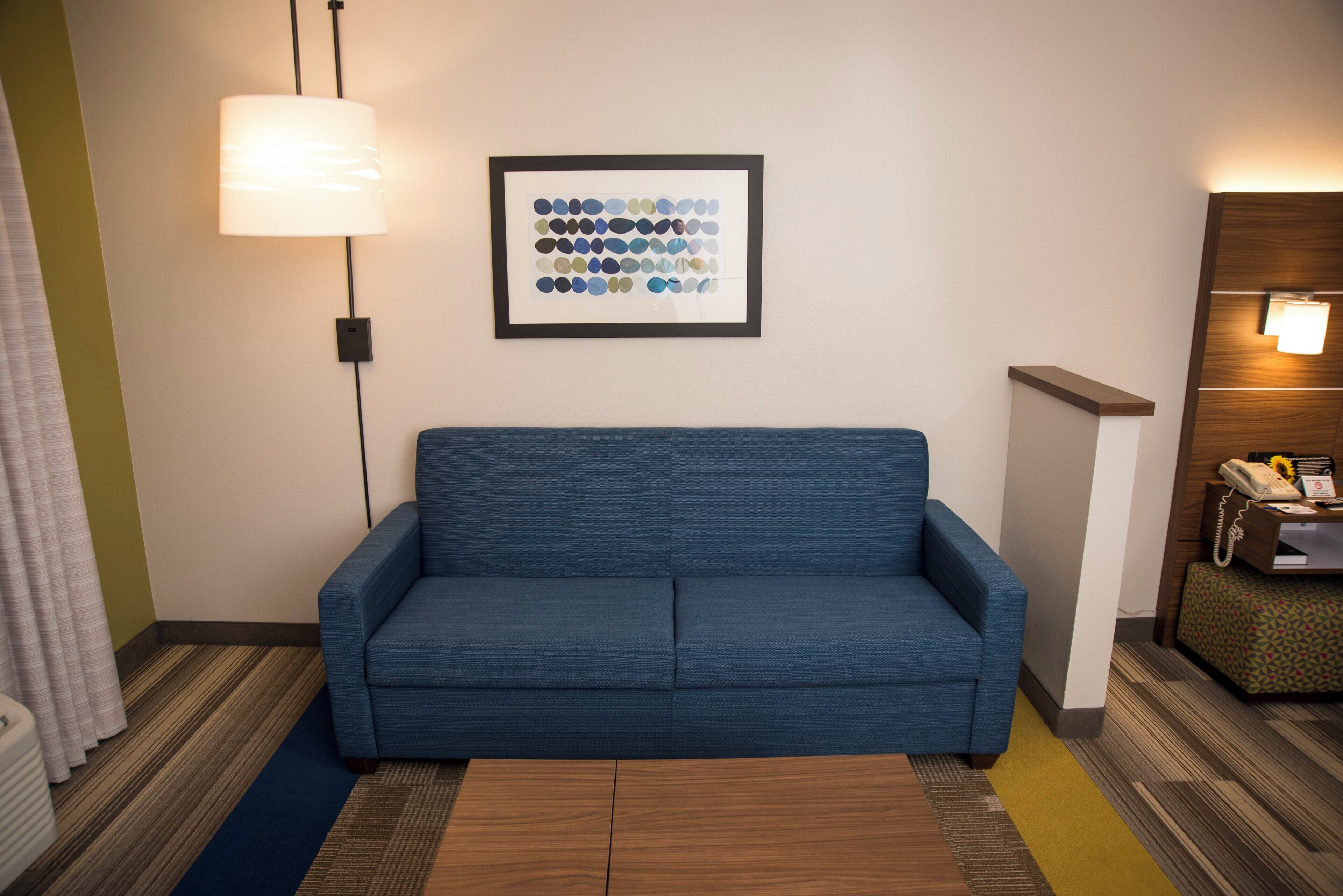 A room with a blue sofa, a framed artwork on the wall, a lamp, and a small desk with a telephone.