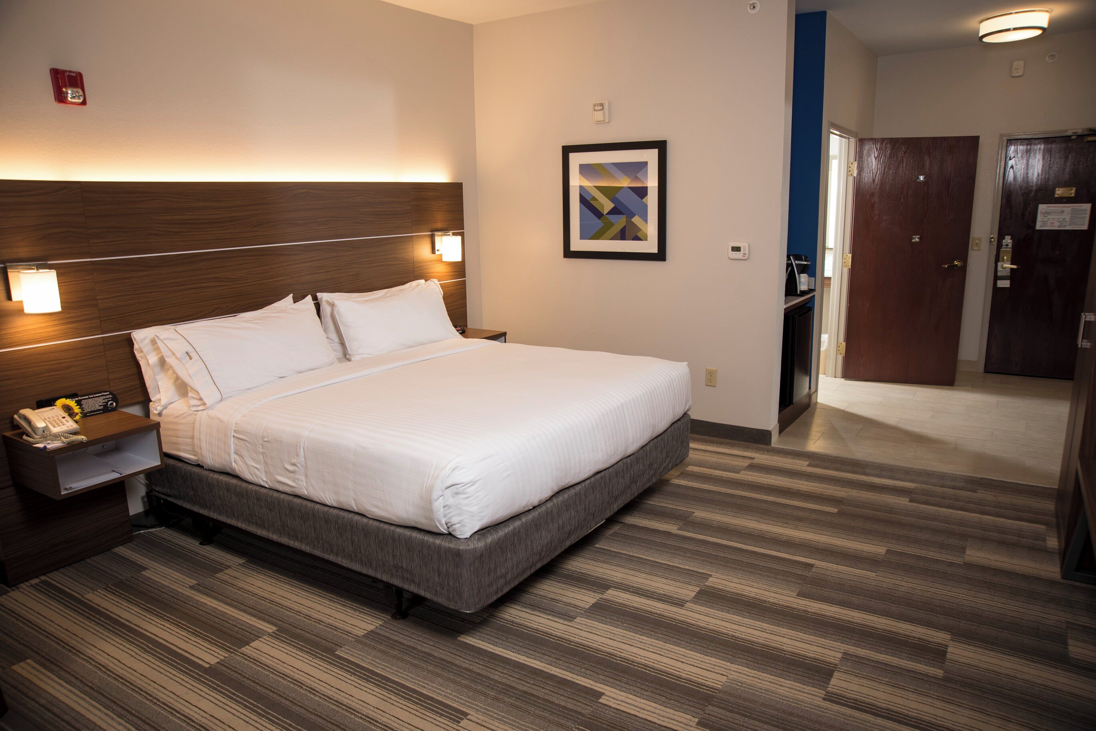 A hotel room with a large bed, bedside table with a lamp, a framed picture on the wall, and a wooden floor.