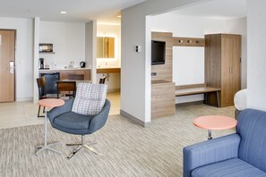 A modern hotel room with a kitchenette, a seating area with a sofa and two round tables, and a TV mounted on the wall.