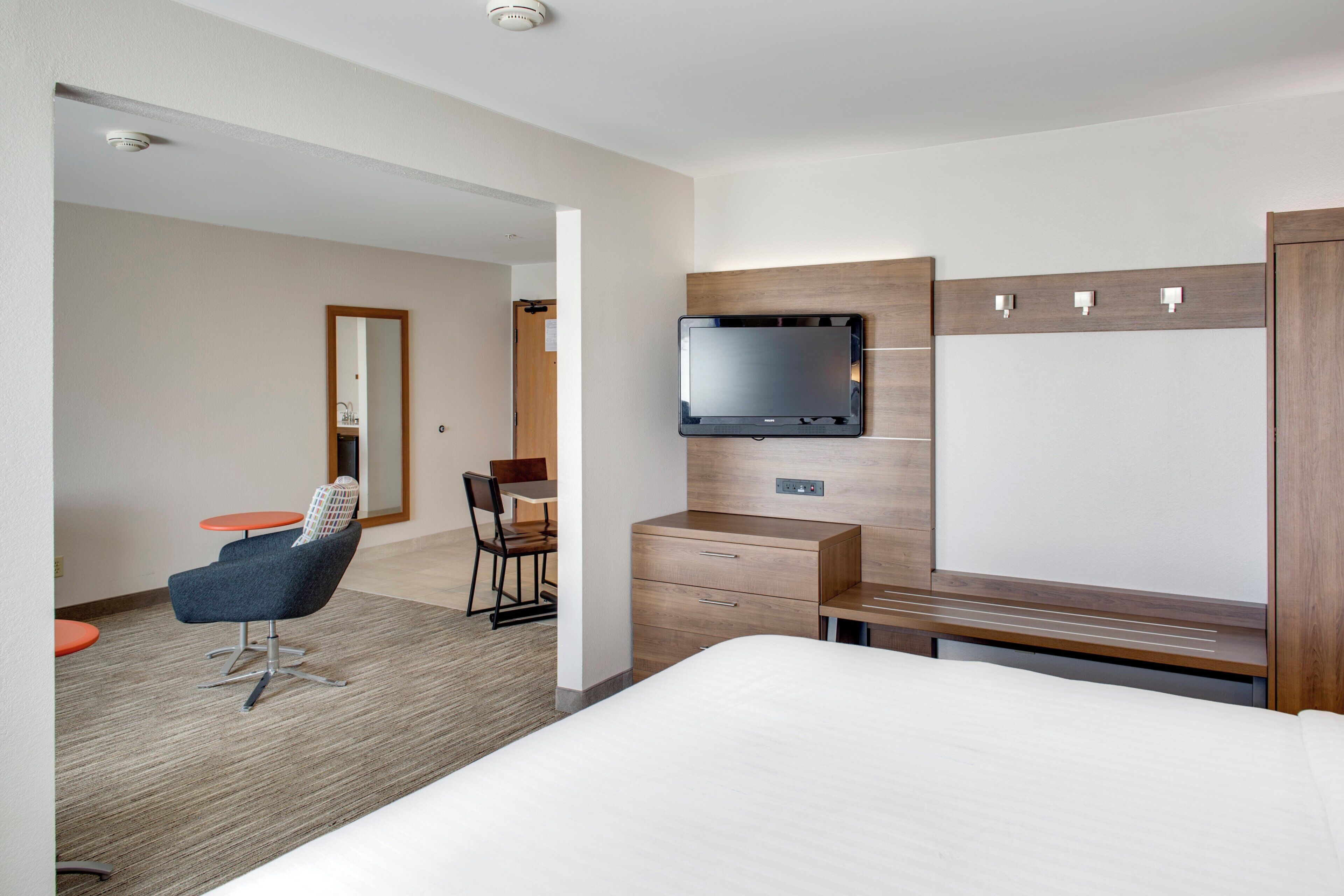 A hotel room with a bed, a desk with a flat-screen TV, a chair, and a small table.