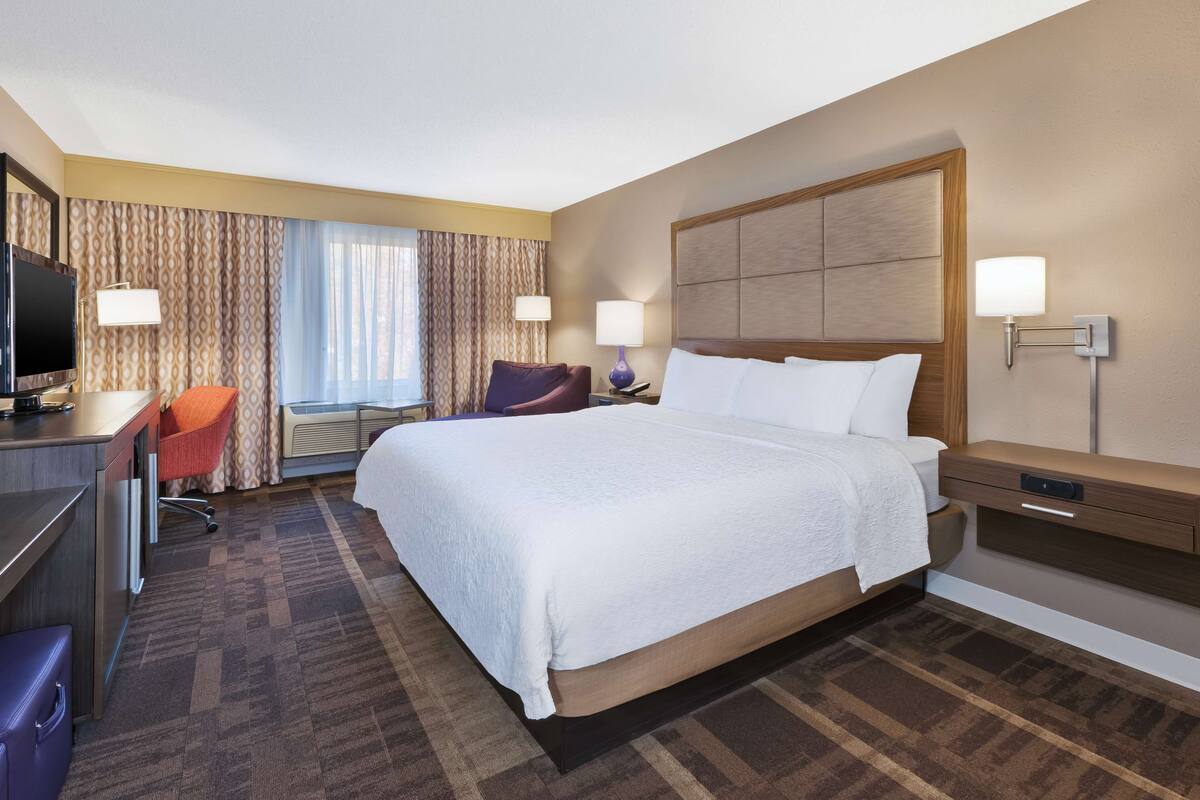 A hotel room with a large bed, a desk with a television, a chair, and a desk lamp.