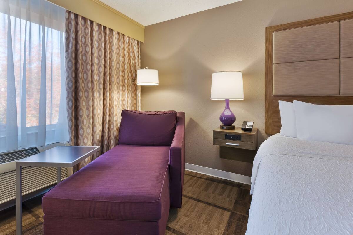 A hotel room with a bed, a sofa, a desk, a lamp, and a window with curtains.
