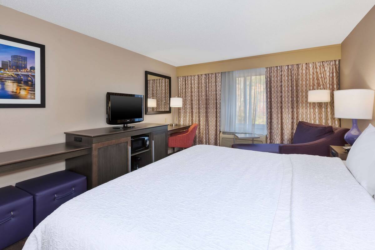 A hotel room with a large bed, a desk with a flat-screen TV, a chair, a sofa, and a window with curtains.