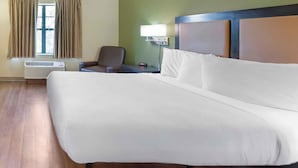 Quarto de hotel com uma cama grande, uma cadeira, uma mesinha de cabeceira com abajur e uma janela com cortinas.